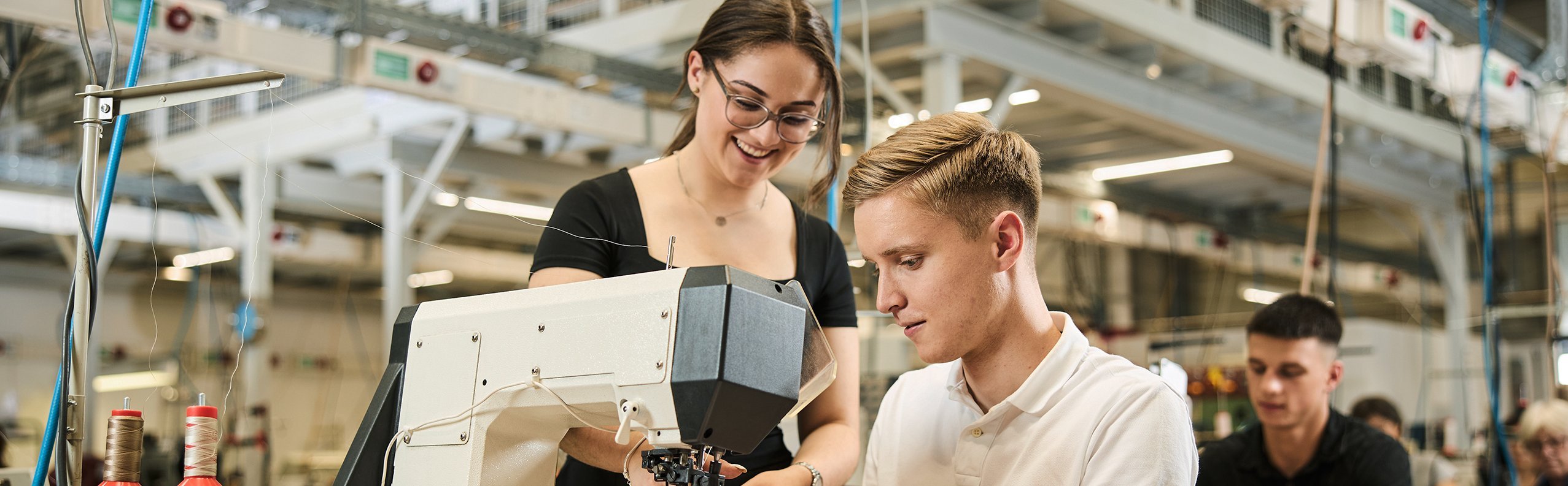 Ein Auszubildender arbeitet in der Produktion an der Nähmaschine. Eine Kollegin steht neben ihm und erklärt etwas.