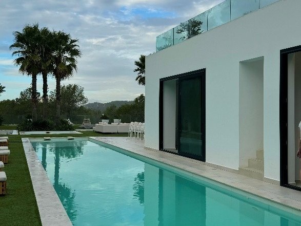 Ein langer türkisfarbener Pool verläuft neben einer weißen Villa. Im Hintergrund sind Palmen zu sehen.