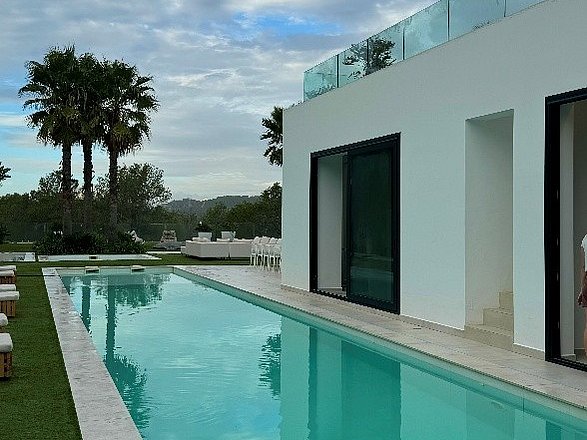 Ein langer türkisfarbener Pool verläuft neben einer weißen Villa. Im Hintergrund sind Palmen zu sehen.