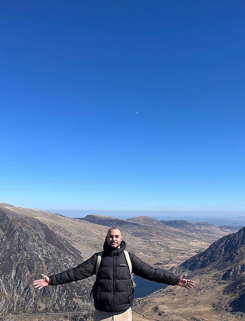 Rieker Auszubildender Jonathan steht mit ausgebreiteten Armen vor einer bergigen Landschaft.