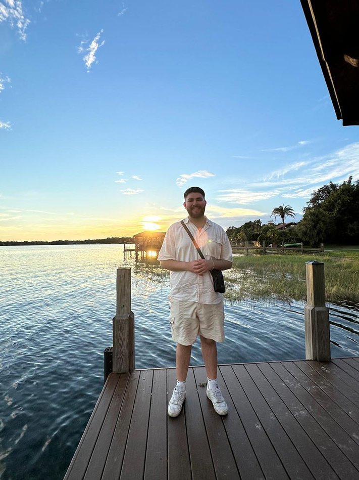 Alpay, dualer Student bei Rieker, auf einem Steg mit Blick auf einen See bei Sonnenuntergang.