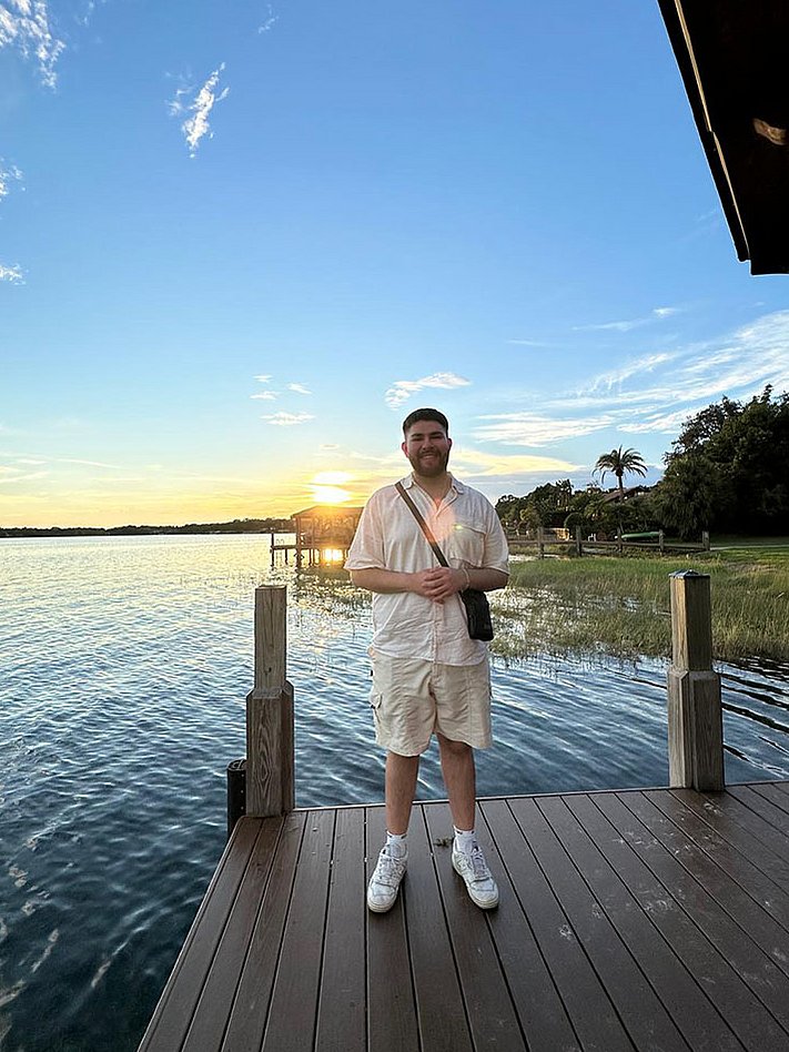 Alpay, dualer Student bei Rieker, auf einem Steg mit Blick auf einen See bei Sonnenuntergang.