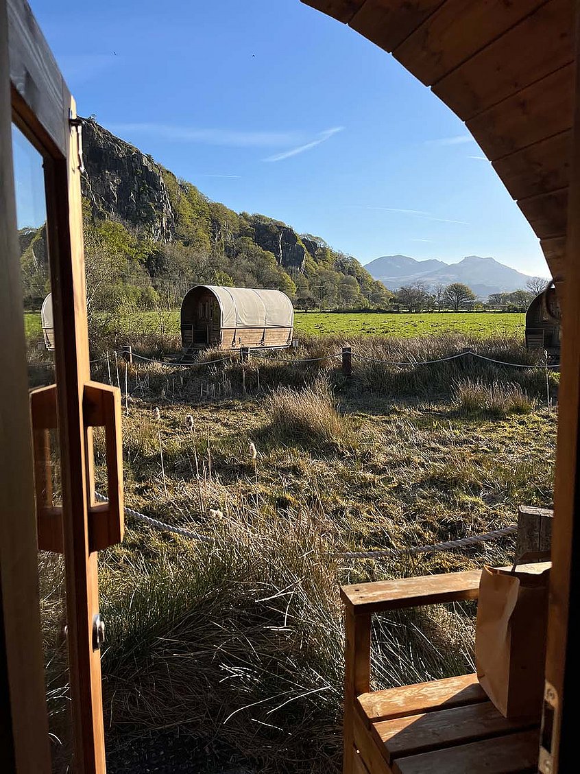 Eine weitläufige Landschaft mit grünen Wiesen, Bergen und blauem Himmel, im Vordergrund steht ein Planwagen.