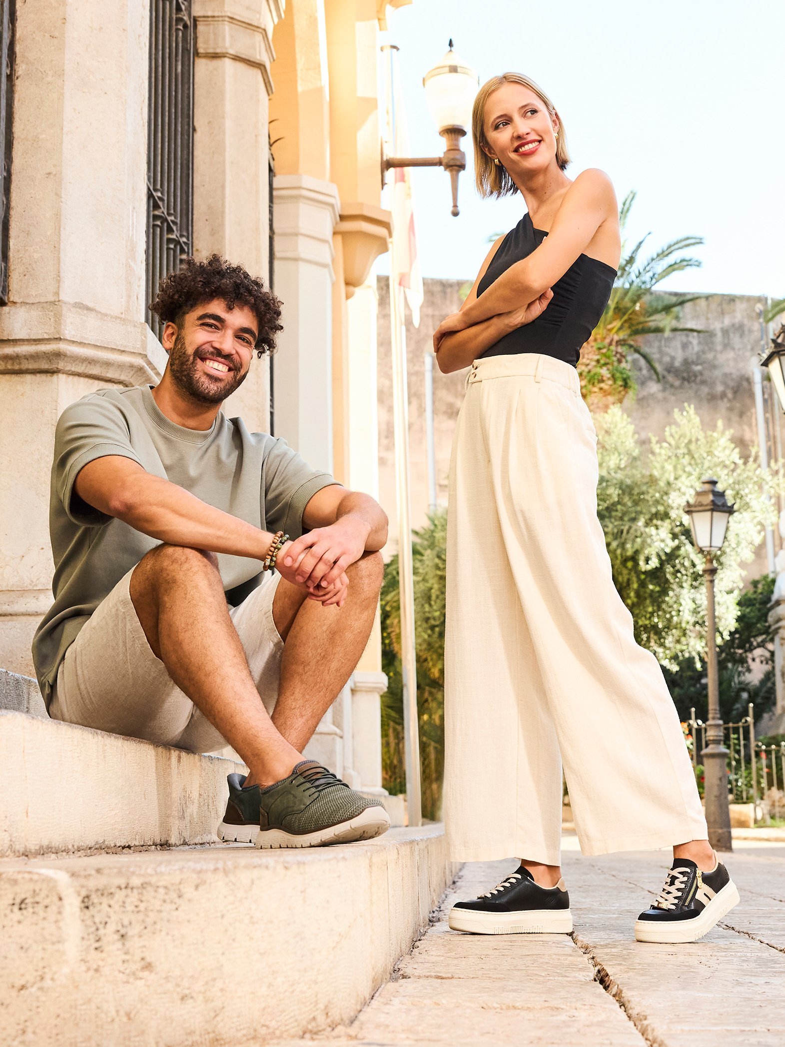 A woman and a man are wearing spring-like everyday outfits with Rieker sneakers and are in a Mediterranean city.