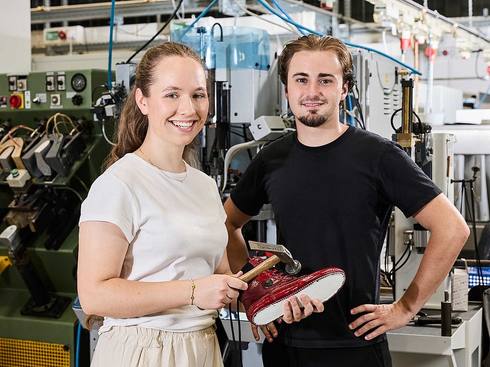 Zwei Auszubildende stehen in der Produktionshalle vor einer Maschine und fügen eine Schuhsohle an einen Schaft.