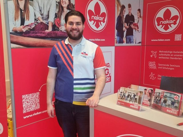Rieker DH Student Alpay steht vor dem Rieker Messestand auf einer Azubimesse.