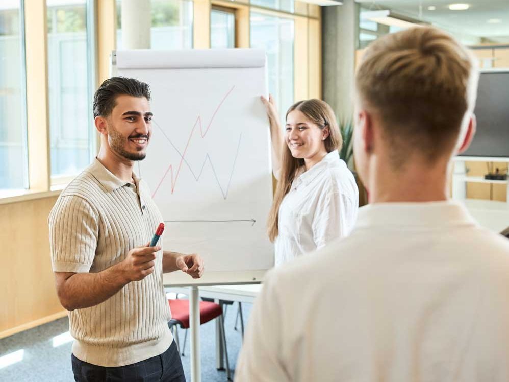 Studierende stehen im Büro vor einem Flip Chart und besprechen ein Diagramm.