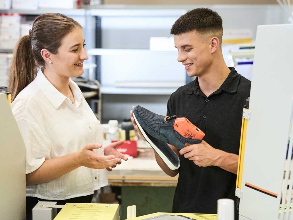 Zwei Auszubildende stehen in der Produktionshalle vor einer Maschine und fügen eine Schuhsohle an einen Schaft. 