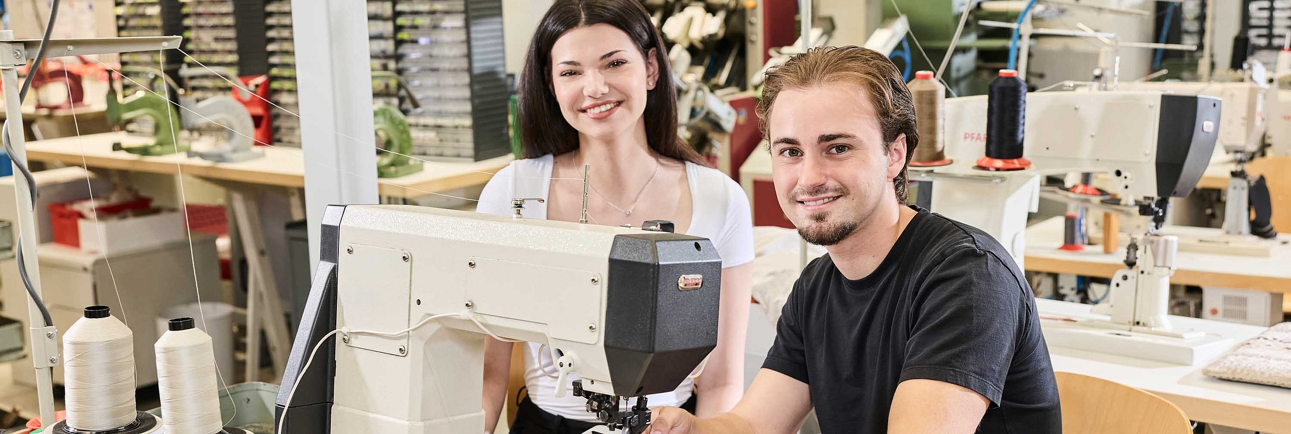 Ein Auszubildender arbeitet in der Produktion an der Nähmaschine. Eine Kollegin steht neben ihm und erklärt etwas.