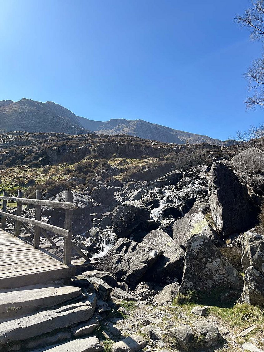 Eine Holzbrücke führt über einen steinigen Bach, im Hintergrund sind Berge vor blauem Himmel.