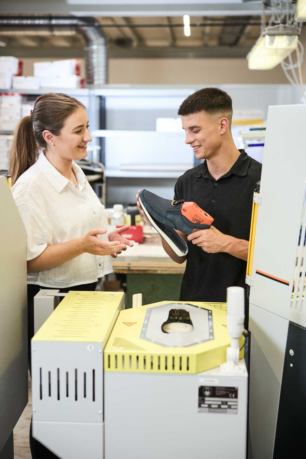 Zwei Auszubildende stehen in der Produktionshalle vor einer Maschine und fügen eine Schuhsohle an einen Schaft.